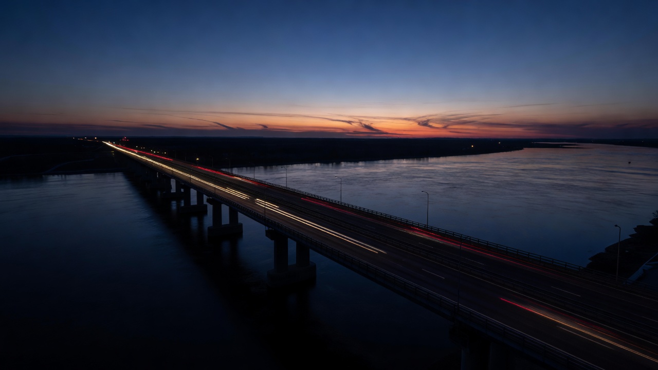 Long road bridge across a wide river at dusk with traffic lights streaking through deep shadow